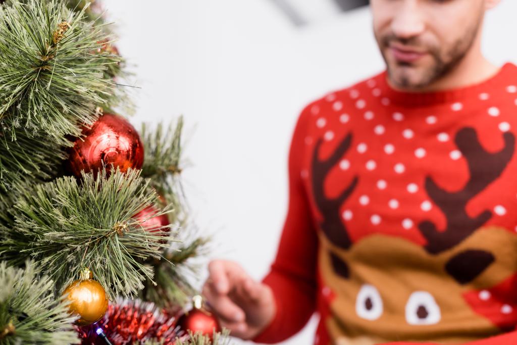 обрезанный вид бородатого мужчины в свитере, украшающего рождественскую елку на размытом переднем плане - Фото, изображение