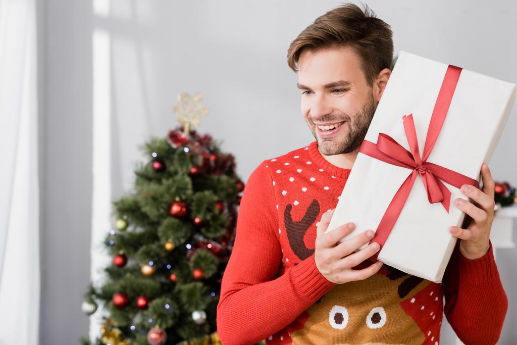 hombre alegre en suéter sosteniendo regalo envuelto cerca del árbol de Navidad sobre fondo borroso - Foto, imagen