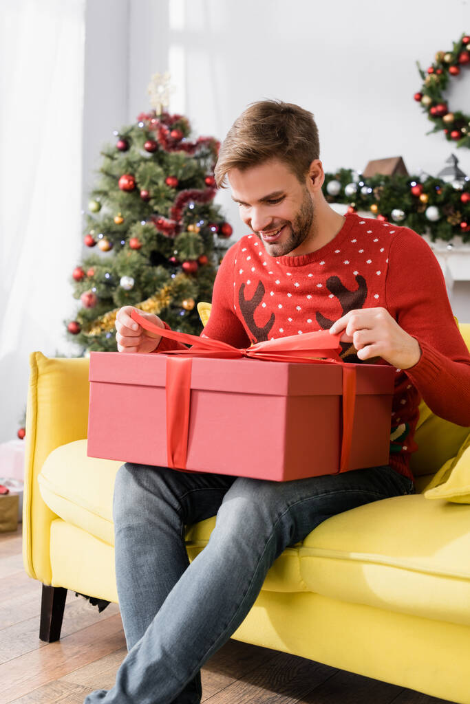 Kırmızı kazaklı mutlu adam arka planda bulanık noel ağacıyla koltukta otururken hediye paketine bakıyor.  - Fotoğraf, Görsel