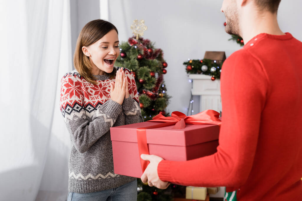 довольный мужчина в красном свитере с завернутым подарком рядом с возбужденной женой и рождественской ёлкой на размытом фоне  - Фото, изображение