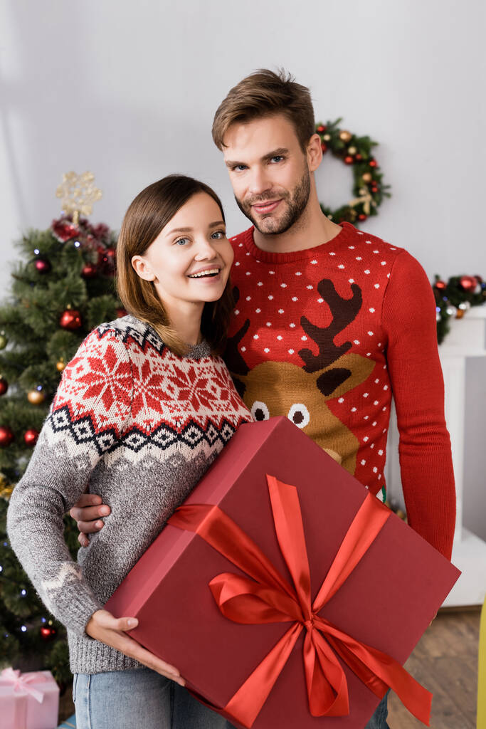 potěšený muž v červeném svetru objímání vzrušený manželka držení dárek v blízkosti vánoční strom na rozmazaném pozadí  - Fotografie, Obrázek