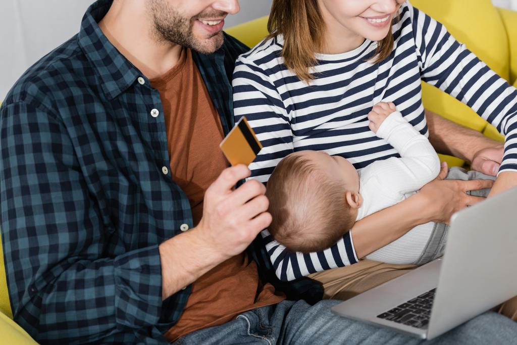 веселый мужчина держит кредитную карту рядом с женой с младенцем сыном и ноутбуком  - Фото, изображение