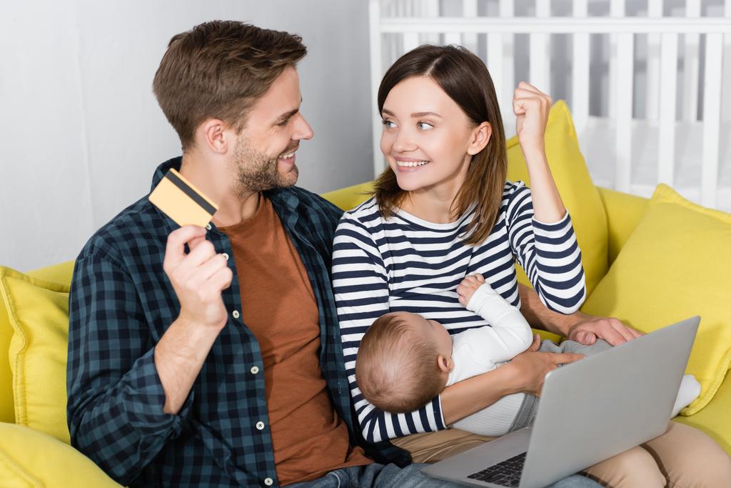 cheerful man holding credit card near excited wife with infant son while online shopping  - Photo, Image