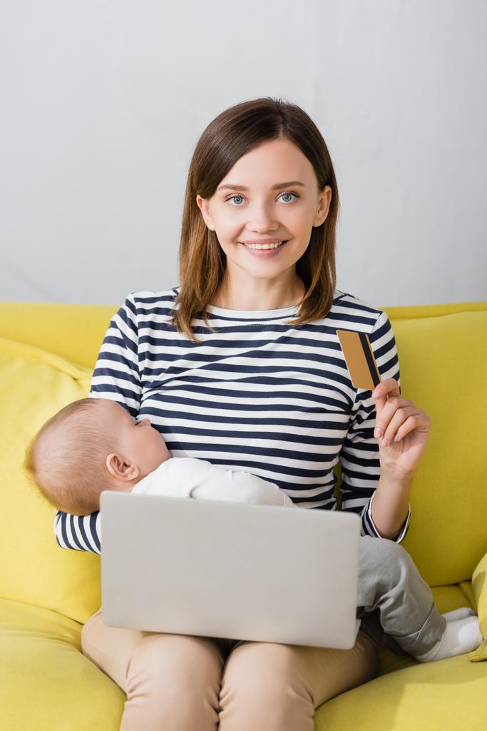 veselá žena s nemluvně syn drží kreditní kartu v blízkosti notebooku na rozmazané popředí  - Fotografie, Obrázek