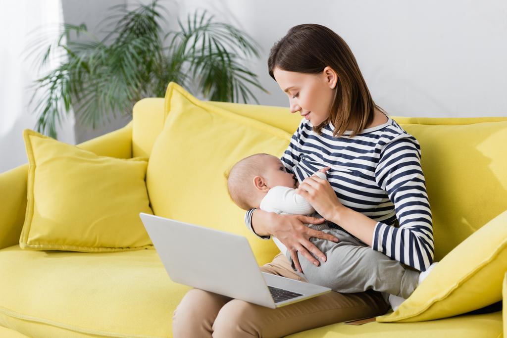 mujer sosteniendo en brazos hijo lactante mientras amamanta cerca de la computadora portátil mientras está sentado en el sofá - Foto, imagen
