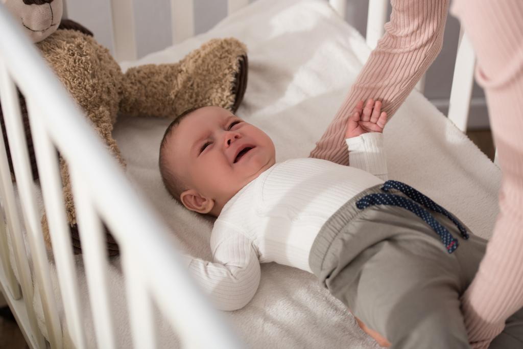 мать поднимает плачущего младенца в кроватке на размытом переднем плане  - Фото, изображение