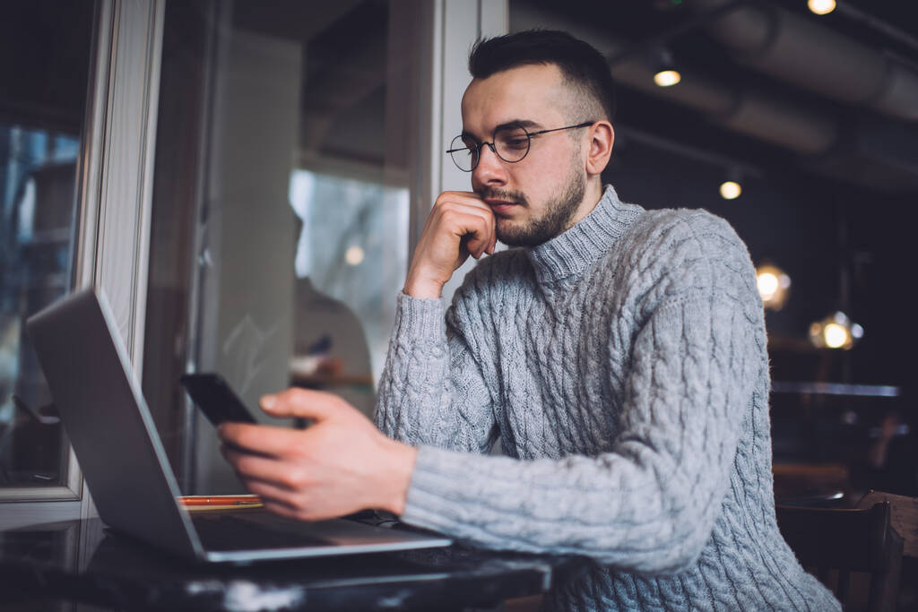Zdola vážný mladý pohledný muž v ležérní oblečení a brýle pomocí smartphonu, zatímco sedí v kavárně u stolu s notebookem opřený o ruce - Fotografie, Obrázek