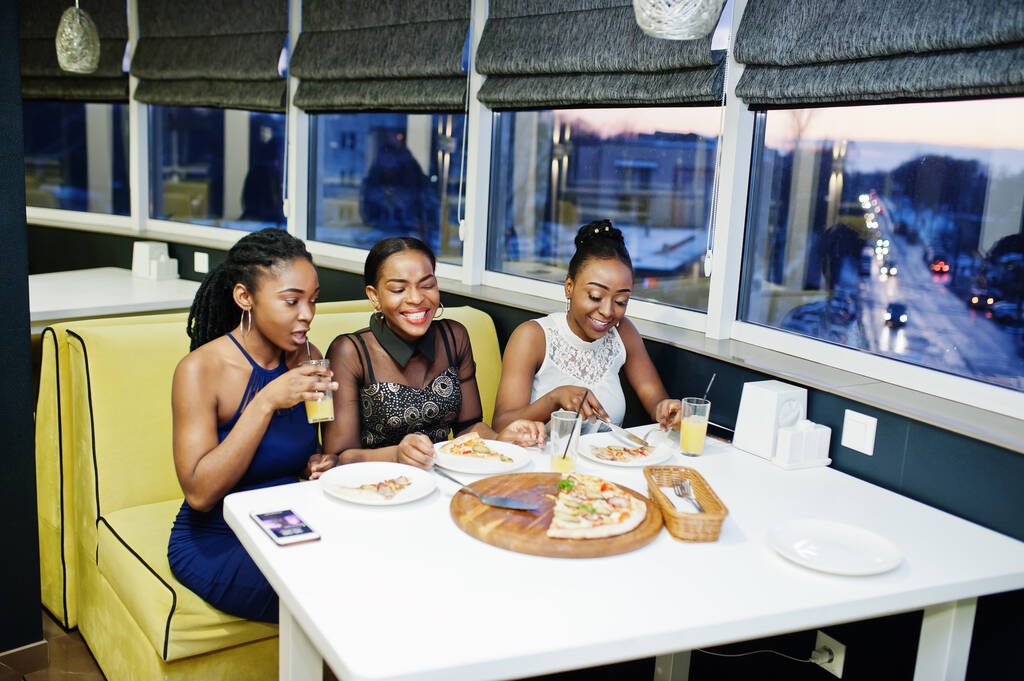 Três africanas de vestido posando no restaurante, comendo pizza e bebendo suco. - Foto, Imagem