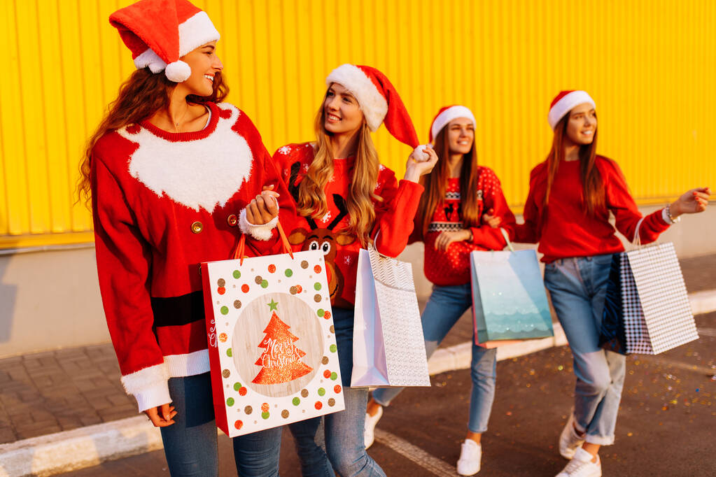 στιλάτες νεαρές γυναίκες, χαρούμενες φίλες με καπέλα santa claus με τσάντες για ψώνια στο φόντο του κίτρινου τοίχου του εμπορικού κέντρου. Ψώνια, Πρωτοχρονιά - Φωτογραφία, εικόνα