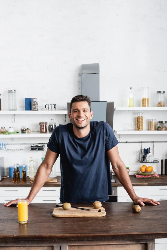 Fiatal férfi mosolyog a kamera közelében kiwi a vágódeszkán és egy pohár narancslé a konyhaasztalon  - Fotó, kép