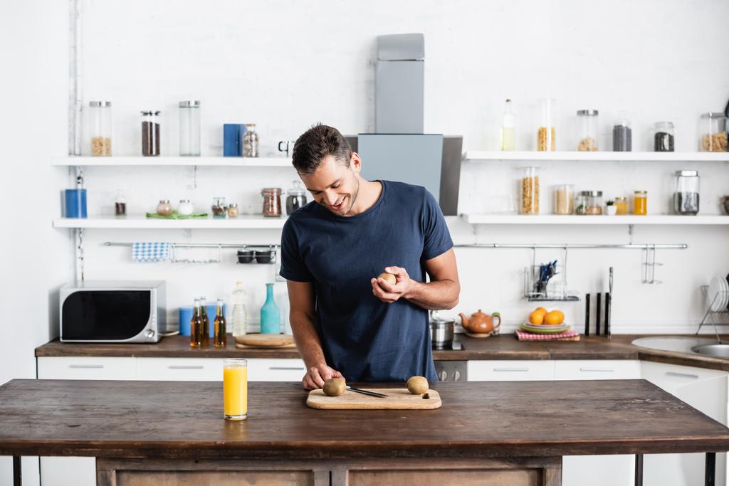 Uśmiechnięty mężczyzna patrzący na kiwi na desce do krojenia w pobliżu soku pomarańczowego w szklance na stole w kuchni  - Zdjęcie, obraz