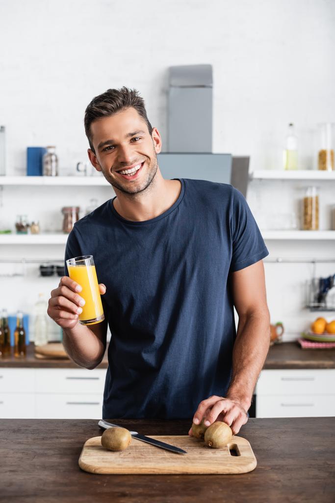 Χαρούμενος άντρας που κρατάει ένα ποτήρι χυμό πορτοκαλιού κοντά στο ακτινίδιο σε ξύλο κοπής στο τραπέζι  - Φωτογραφία, εικόνα