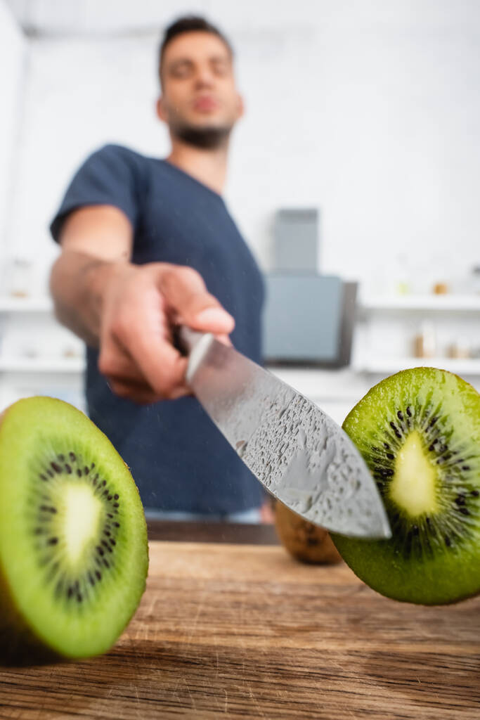Vista da vicino di metà del succoso kiwi vicino al coltello in gocce d'acqua in mano all'uomo su sfondo sfocato  - Foto, immagini
