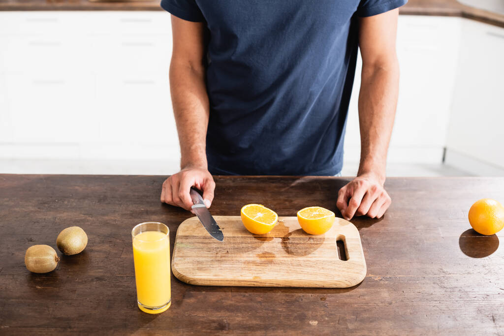 Cropped view of man holding knife near oranges, kiwi and glass of orange juice on kitchen table  - Foto, Imagen