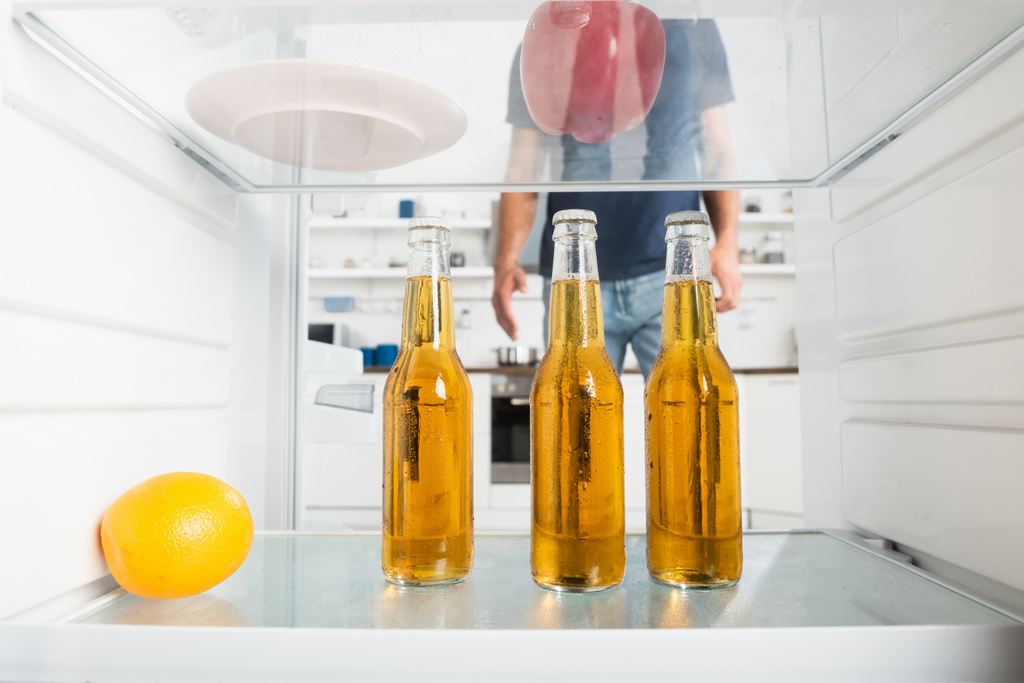 Бутылки пива и апельсин в холодильнике рядом с человеком на размытом фоне  - Фото, изображение