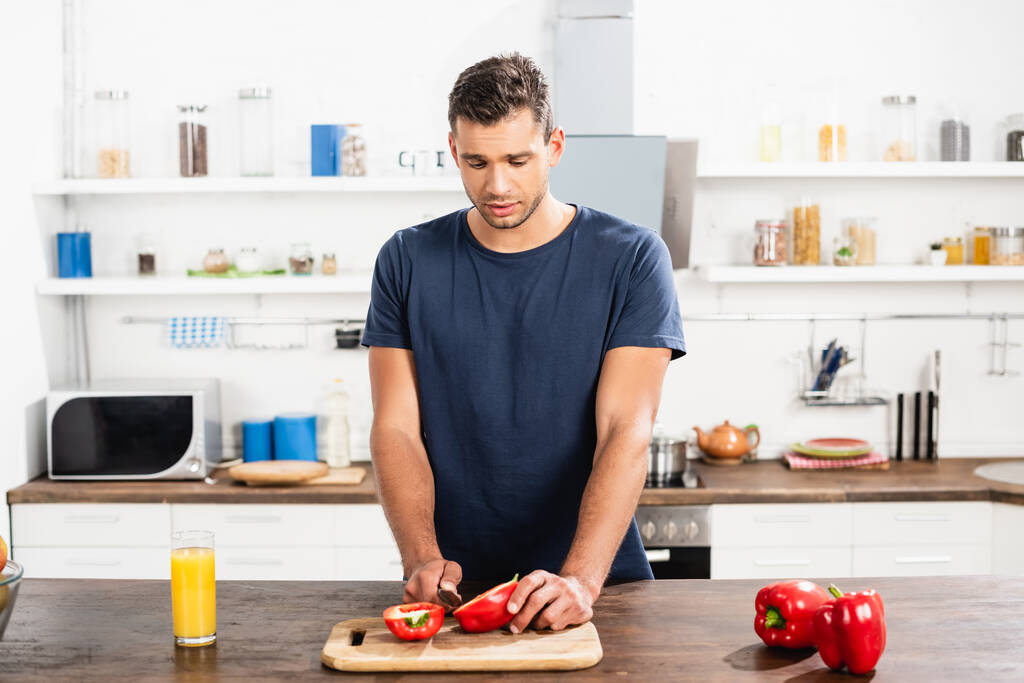 Junger Mann schneidet Paprika neben Glas Orangensaft in Küche  - Foto, Bild