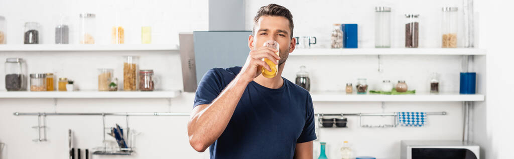 Junger Mann schaut in die Kamera, während er Orangensaft in der Küche trinkt, Banner  - Foto, Bild