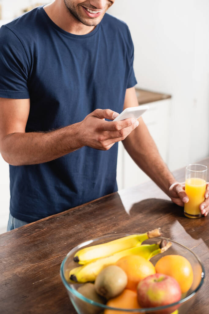 オレンジジュースのガラスの近くでスマートフォンを使用して笑顔の男とぼやけた前景に新鮮な果物の作物のビュー  - 写真・画像