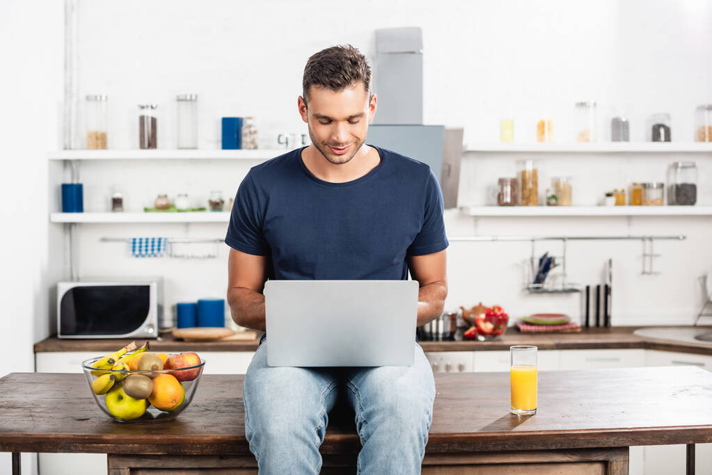 Młody człowiek korzystający z laptopa przy szklance soku pomarańczowego i świeżych owoców w kuchni  - Zdjęcie, obraz