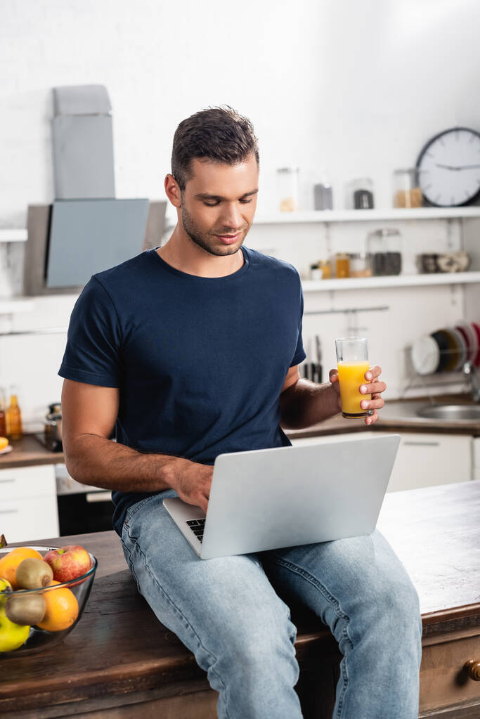 Mann mit Laptop und Orangensaft sitzt neben Obst auf Küchentisch  - Foto, Bild