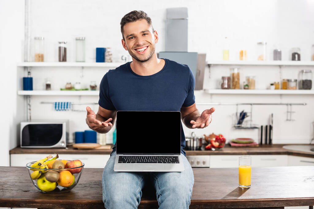 Vidám férfi mutató kezek közelében laptop üres képernyő, gyümölcsök és narancslé a konyhaasztalon  - Fotó, kép
