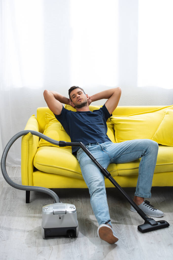 Young man sitting on couch near vacuum cleaner on blurred foreground - Foto, Imagen