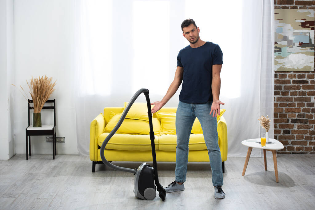Young man pointing with hands near vacuum cleaner in living room  - Photo, Image