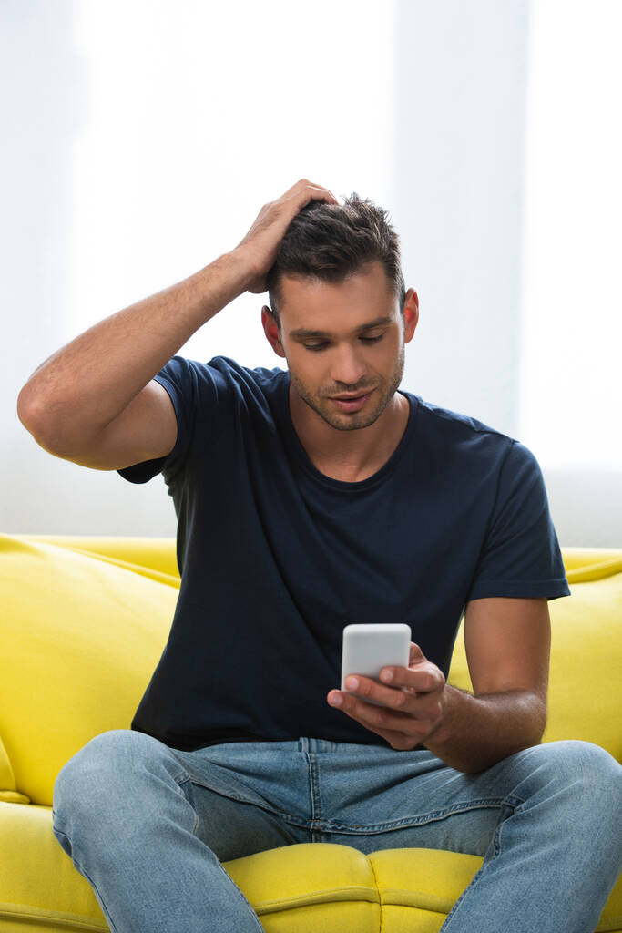 Uomo con mano vicino alla testa utilizzando smartphone su divano giallo  - Foto, immagini