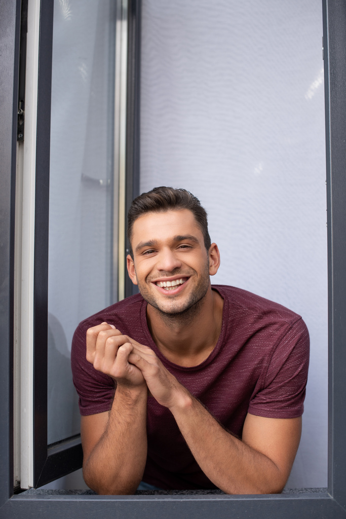 Iloinen mies hymyilee kameralle lähellä avointa ikkunaa kotona  - Valokuva, kuva