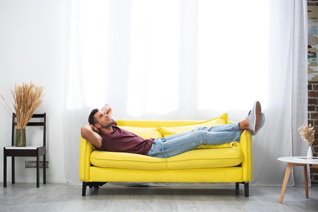 Улыбающийся мужчина с руками возле головы, лежащими дома на диване  - Фото, изображение