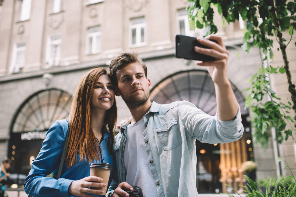 Klidný dospělý muž a atraktivní usmívající se žena s jít teplý drink přičemž selfie s chytrým telefonem, zatímco stojí na ulici proti rozmazané fasády staré budovy ve dne - Fotografie, Obrázek