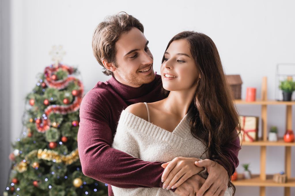 glückliches Paar in warmen Pullovern, das sich in der Nähe des geschmückten Weihnachtsbaums auf verschwommenem Hintergrund umarmt - Foto, Bild