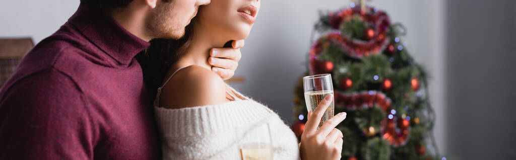 vista recortada de hombre apasionado besar mujer y celebración de copas con champán cerca de árbol de Navidad sobre fondo borroso, pancarta - Foto, Imagen
