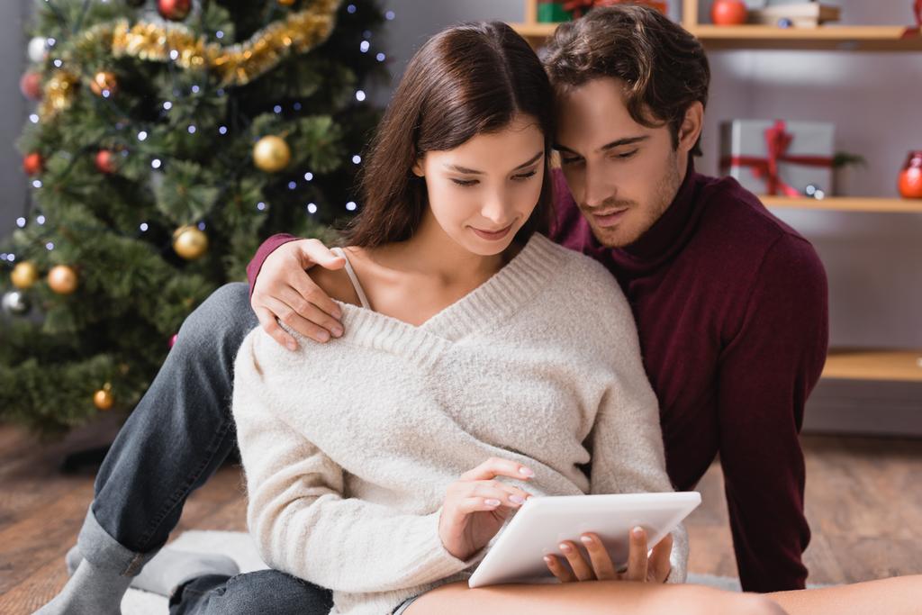 クリスマスツリーの近くにあるデジタルタブレットを背景にセーターを着たカップルが - 写真・画像