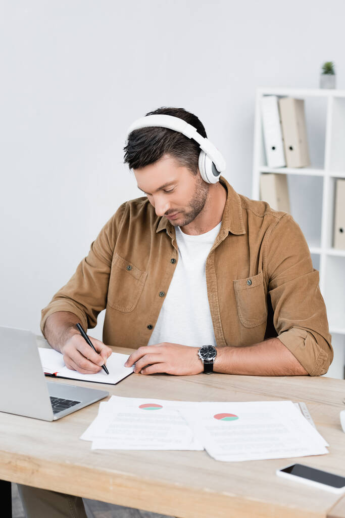 Geschäftsmann mit Kopfhörer schreibt in Notizbuch, während er am Arbeitsplatz mit digitalen Geräten sitzt - Foto, Bild