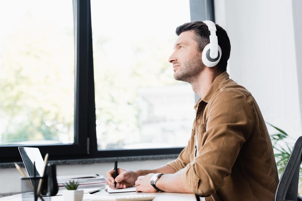 Vista lateral del hombre de negocios en auriculares con lápiz mirando hacia otro lado mientras está sentado en el lugar de trabajo con ventana borrosa en el fondo - Foto, Imagen