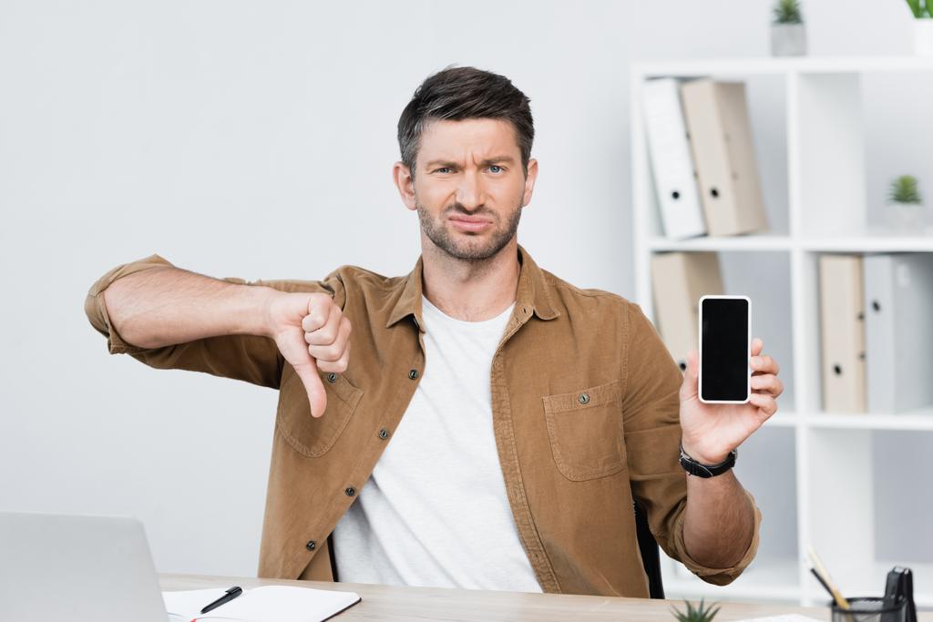 Незадоволений бізнесмен з великим пальцем вниз, дивлячись на камеру, тримаючи смартфон на робочому місці на розмитому фоні
 - Фото, зображення