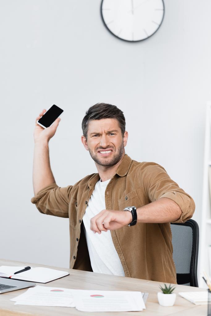 職場でスマートフォンを取り払いながらカメラを見ている怒っているビジネスマン - 写真・画像