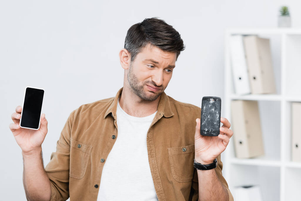 Ontevreden zakenman met smartphone op zoek naar kapotte mobiele telefoon op wazige achtergrond - Foto, afbeelding