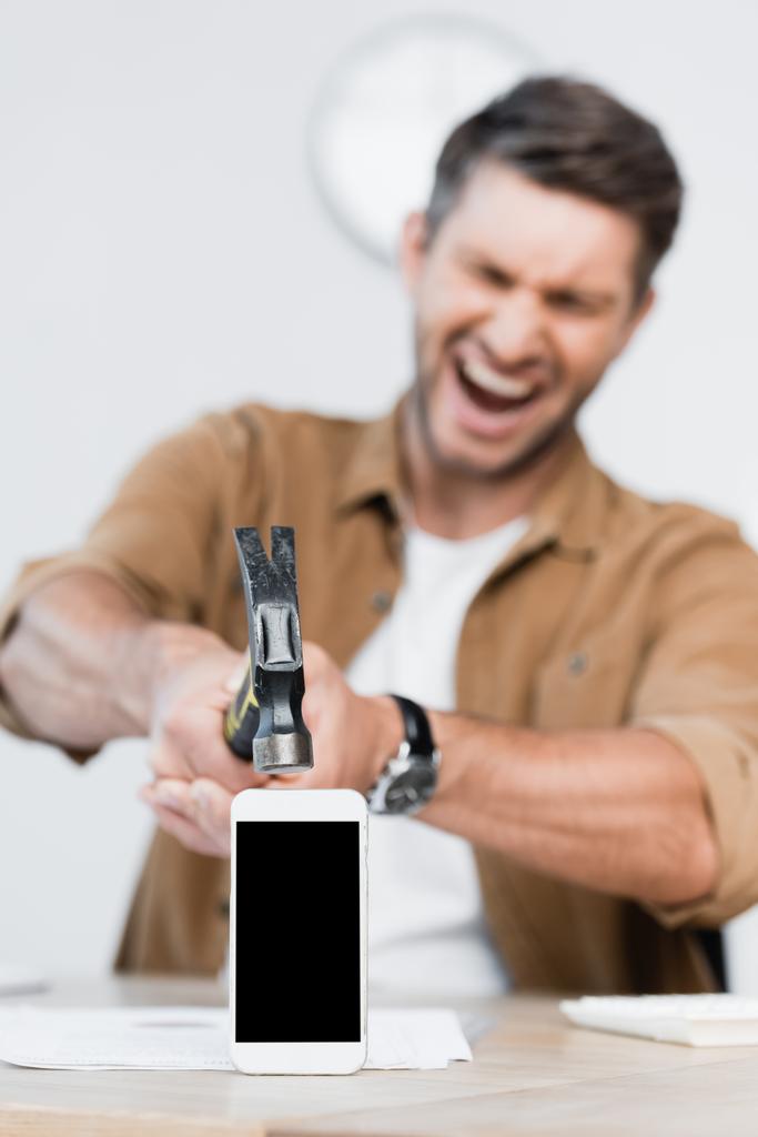 Aggressivo uomo d'affari gridando tenendo martello vicino smartphone con schermo bianco sul tavolo su sfondo sfocato - Foto, immagini