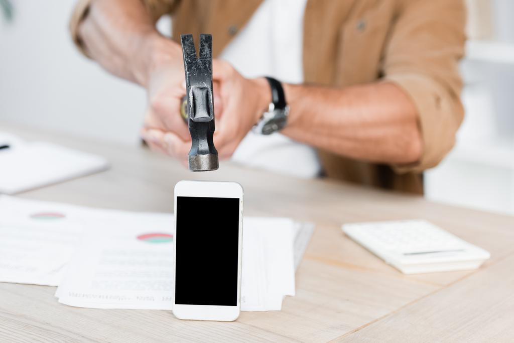 Vista cortada do empresário segurando martelo perto do smartphone com tela em branco no local de trabalho no fundo embaçado - Foto, Imagem