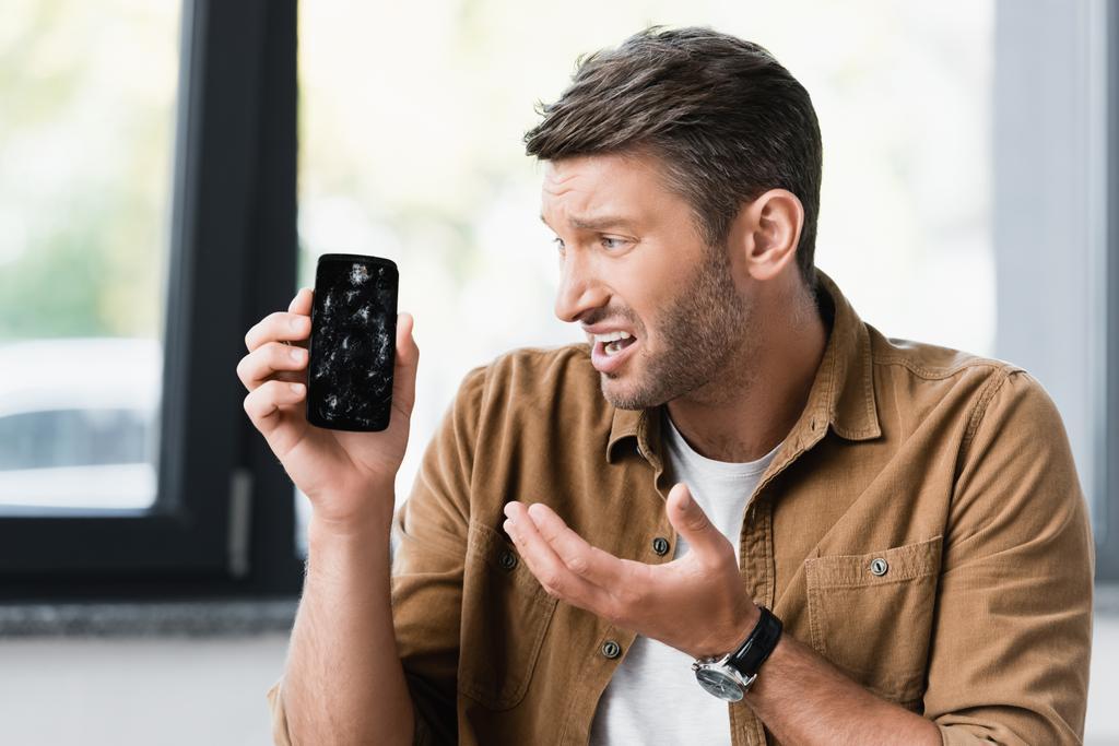 Vyděšený podnikatel při pohledu na rozbitý smartphone na rozmazaném pozadí - Fotografie, Obrázek