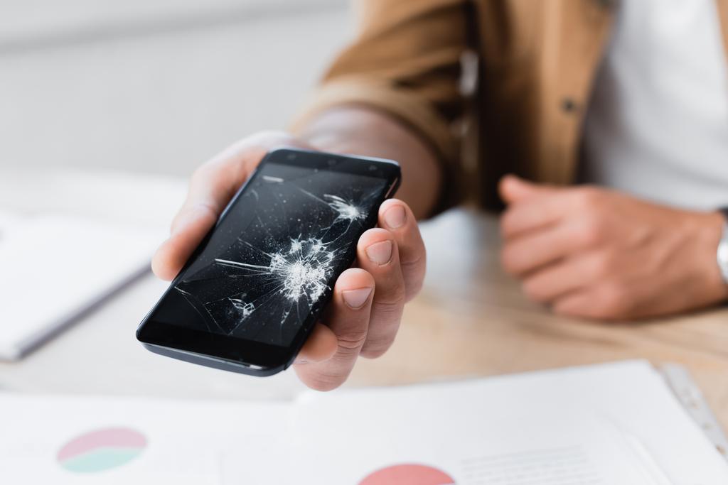 Oříznutý pohled na muže držícího rozbitý smartphone s rozmazaným pracovištěm na pozadí - Fotografie, Obrázek