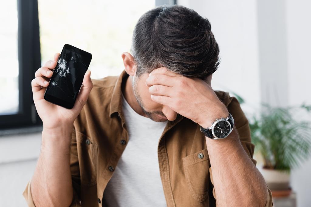 Disappointed businessman with hand on forehead, holding smashed smartphone on blurred background - Photo, Image
