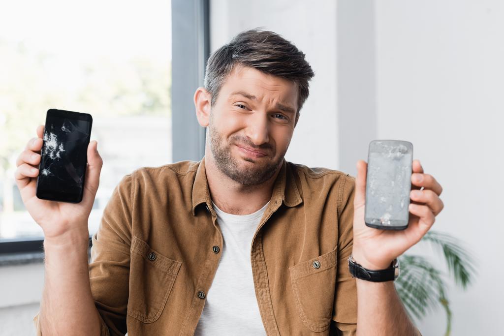 Hombre de negocios confundido con gesto de encogimiento de hombros mirando a la cámara mientras sostiene teléfonos inteligentes aplastados sobre un fondo borroso - Foto, imagen