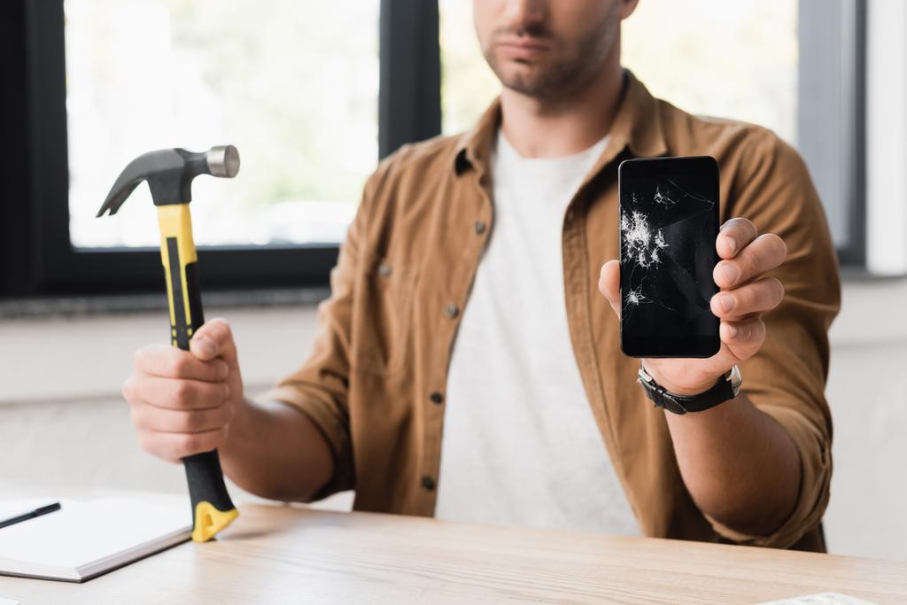 Ausgeschnittene Ansicht eines Geschäftsmannes mit Hammer, der beschädigtes Smartphone zeigt, während er am Arbeitsplatz auf verschwommenem Hintergrund sitzt - Foto, Bild