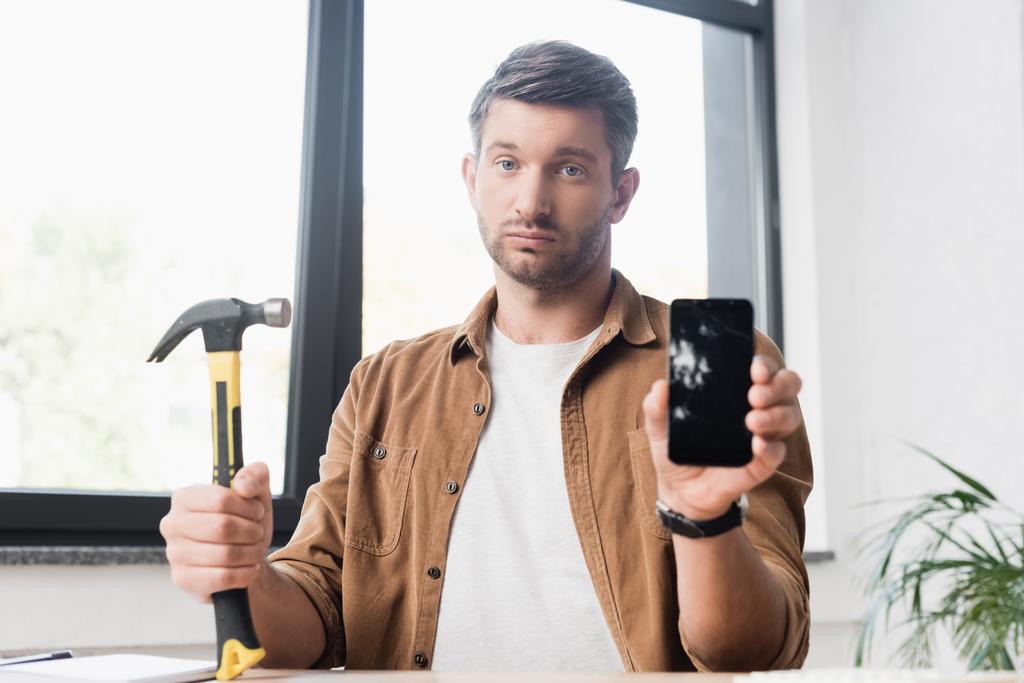 Trauriger Geschäftsmann mit Hammer blickt in Kamera und zeigt beschädigtes Smartphone mit verschwommenem Fenster im Hintergrund - Foto, Bild