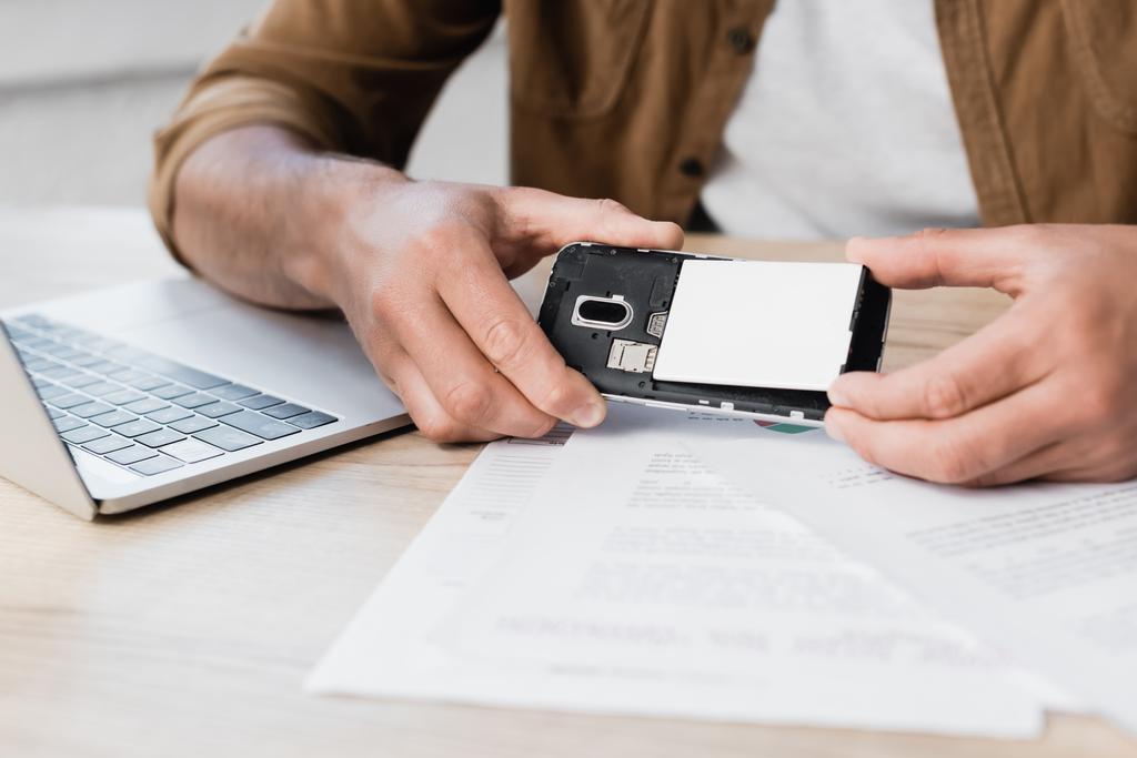 Обрезанный вид бизнесмена, помещающего батарейки в мобильный телефон на рабочем месте с бумажной работой на размытом переднем плане - Фото, изображение