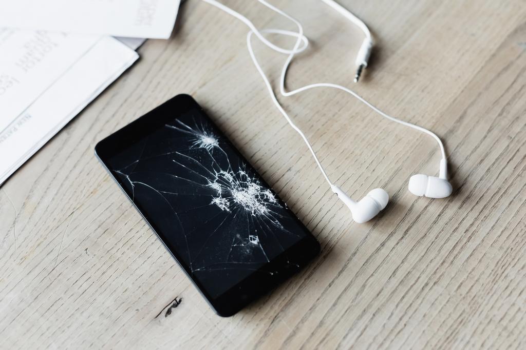 Detailní pohled na rozbitý mobil se sluchátky a papírovými listy na dřevěném pozadí - Fotografie, Obrázek