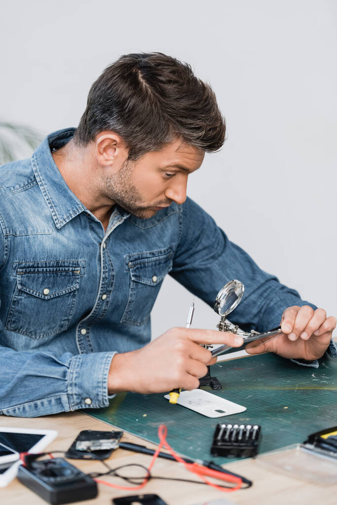Reparaturarbeiter mit Schraubenzieher blickt durch Lupe auf zerlegtes Teil des Mobiltelefons am Tisch auf verschwommenem Vordergrund - Foto, Bild